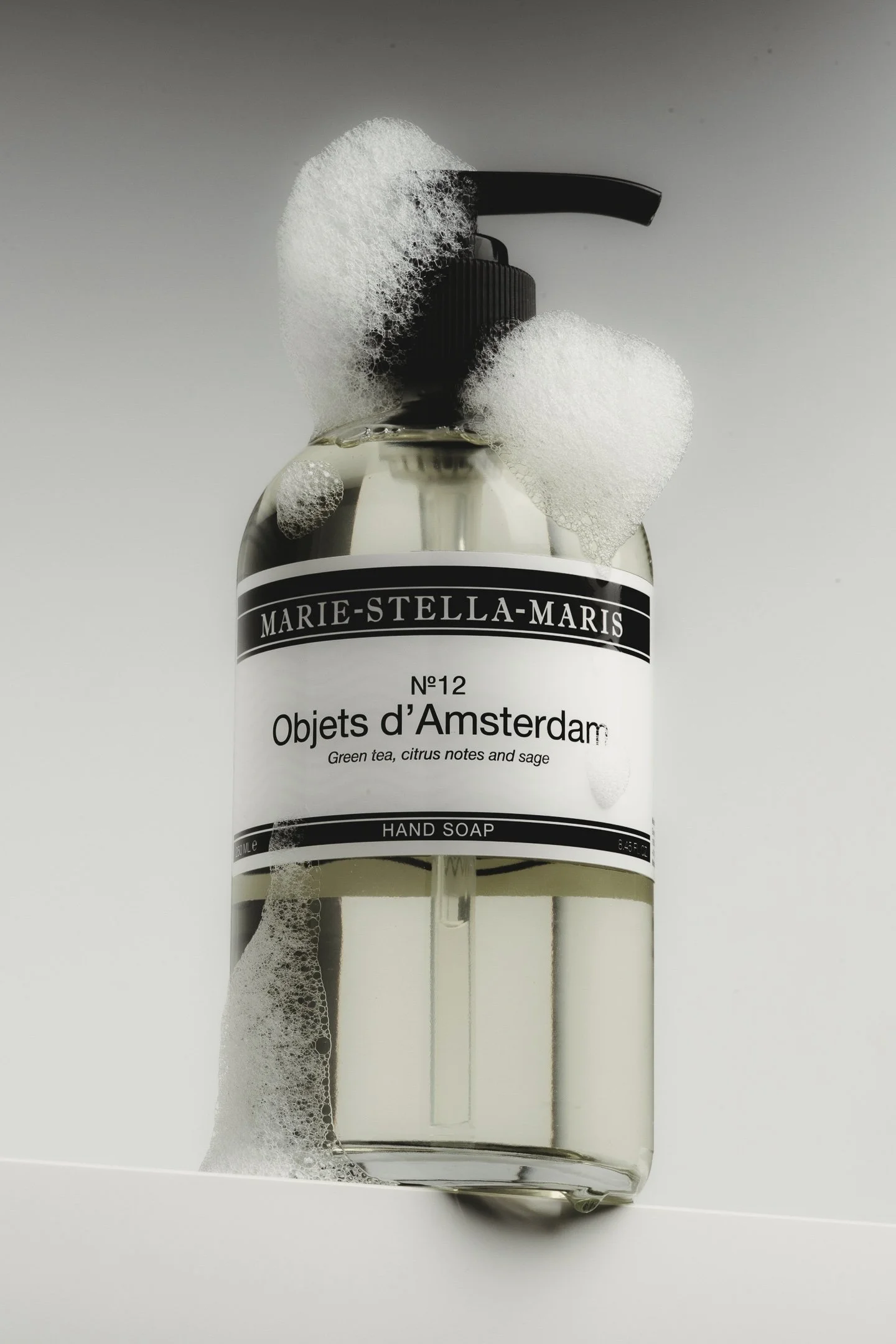 A clear plastic bottle of Marie-Stella-Maris hand soap with a black pump dispenser. The soap is labeled as "Objets d'Amsterdam" with a scent of green tea, citrus notes, and sage. The bottle is covered in soap bubbles.