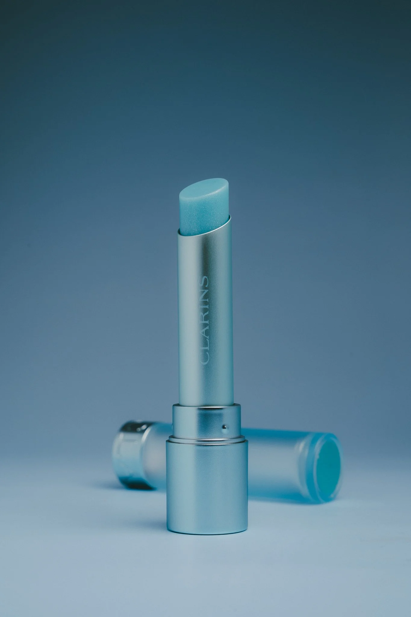 A close-up of a skincare product tube with a light blue cap standing upright against a matching blue background, with other similar tubes lying nearby.