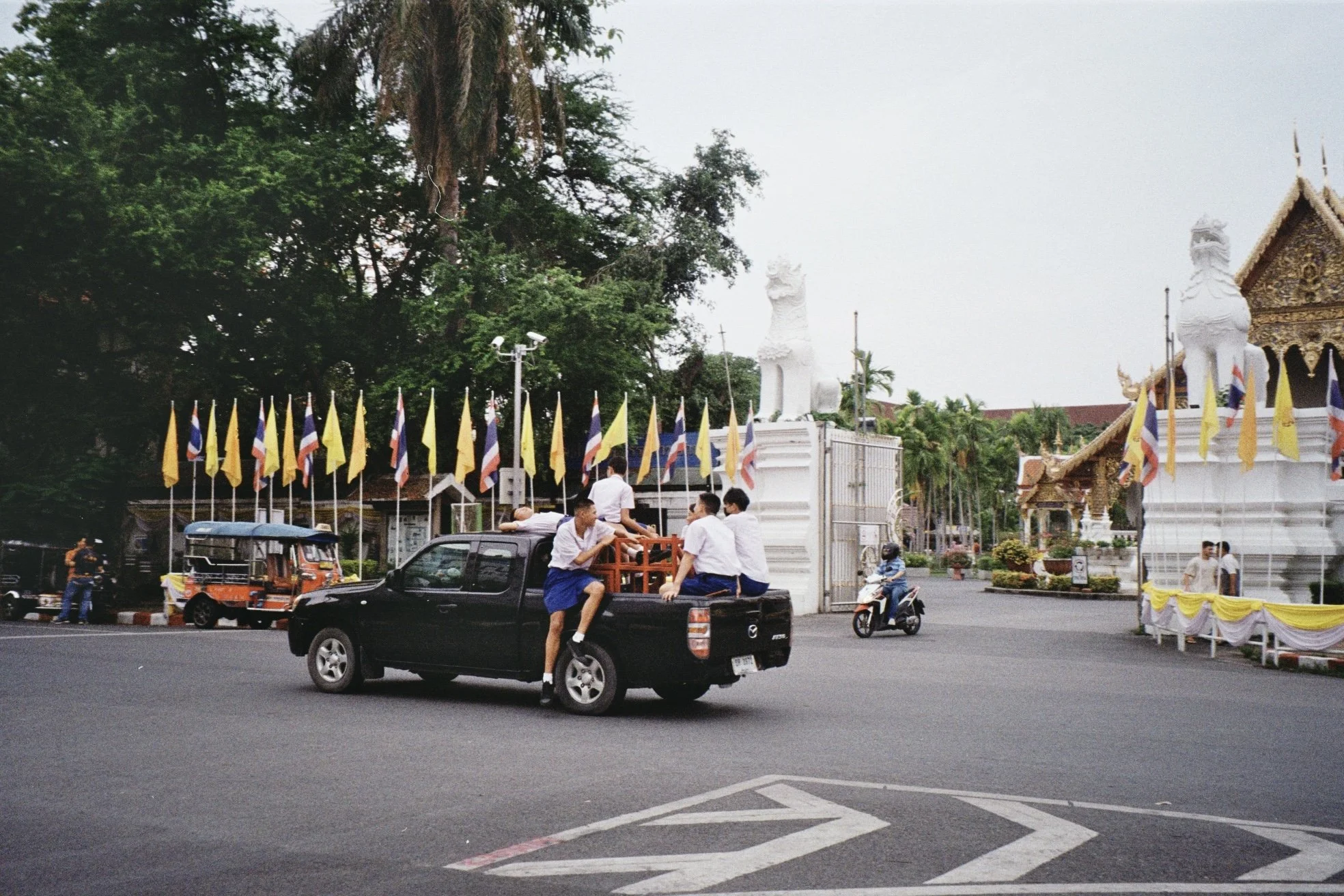 People gathered in front of a temple or religious structure decorated with flags, with large white statues of mythical creatures, trees, and a cloudy sky in the background.