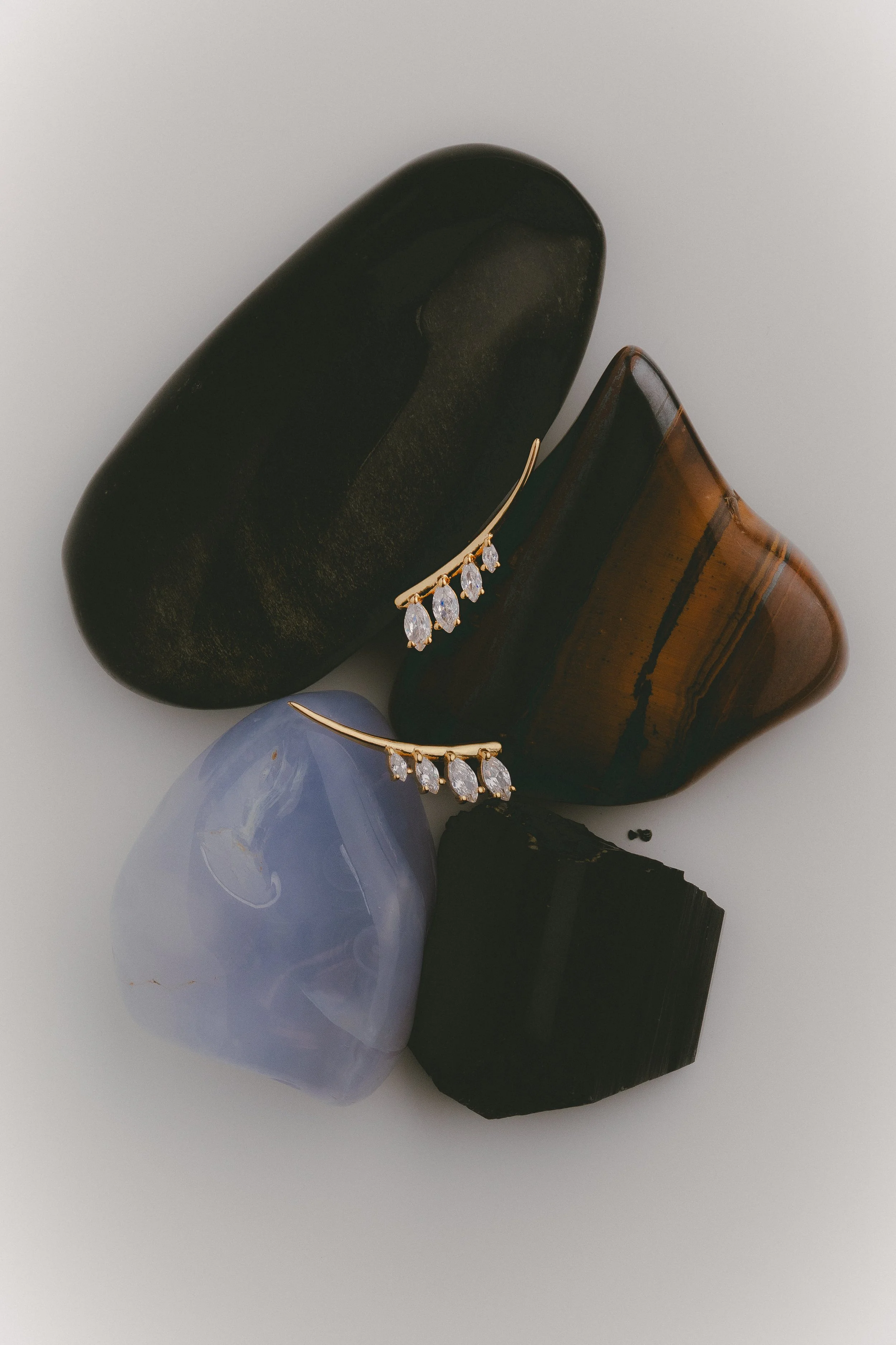 Close-up of a pair of gold earrings with teardrop-shaped clear stones, resting on smooth, rounded stones of black, brown, and blue colors.