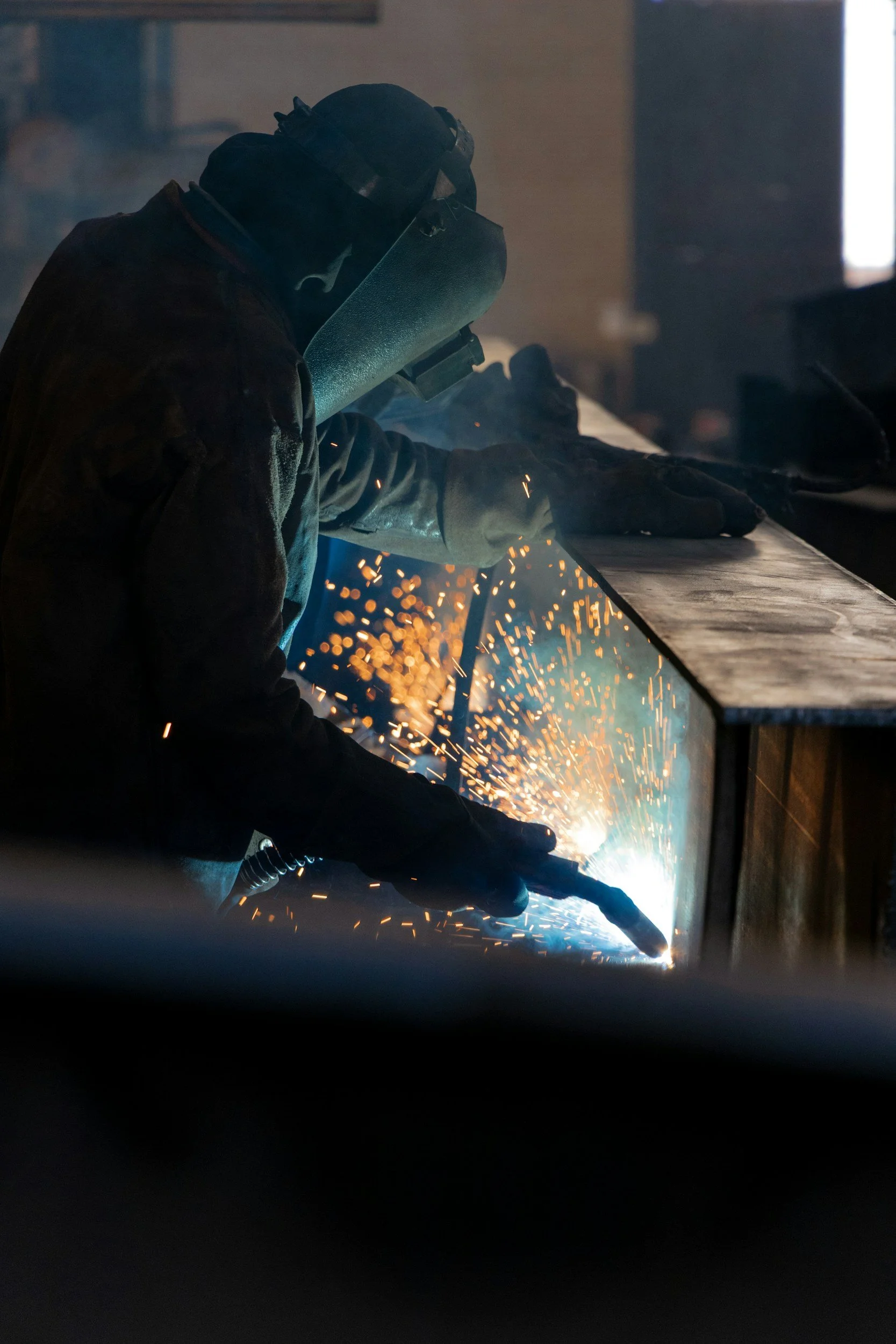 A person welding metal with sparks flying, wearing protective gear including a helmet and gloves, in an industrial workshop.