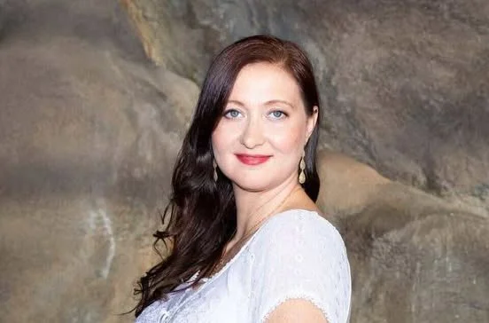 A woman with long dark hair and blue eyes, wearing a white top, standing outdoors against a rocky background.