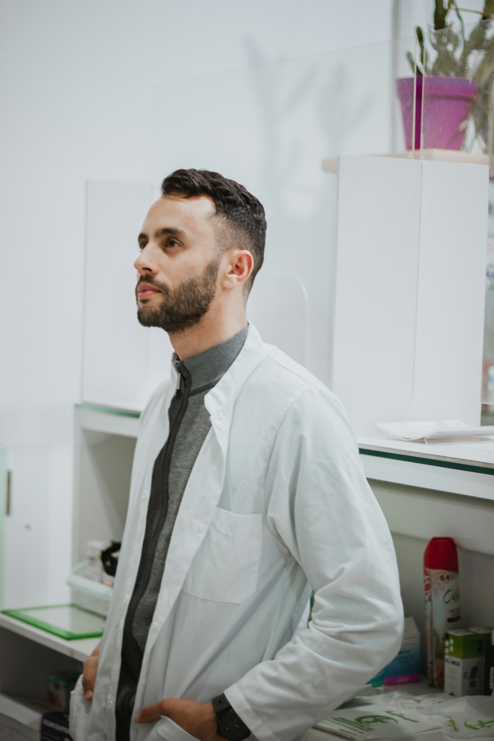 A man with short dark hair and a beard wearing a gray shirt and white lab coat, standing in a laboratory with shelves of supplies and a purple plant pot in the background.
