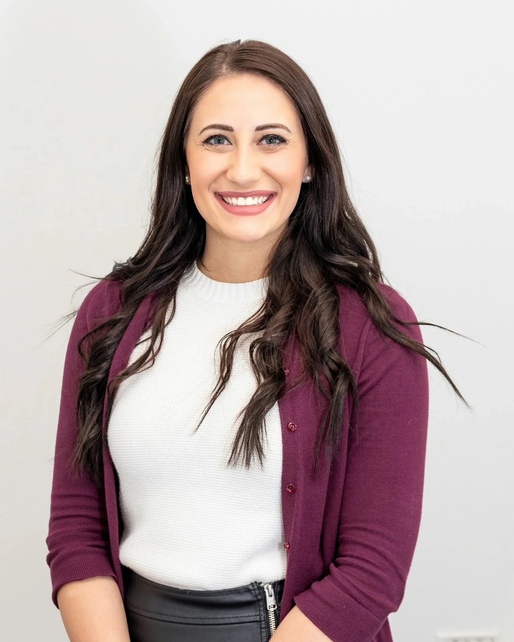 A smiling woman with long dark brown hair, blue eyes, and earrings standing against a plain white background. She is wearing a white top, a magenta cardigan, and a black leather skirt with a zipper.