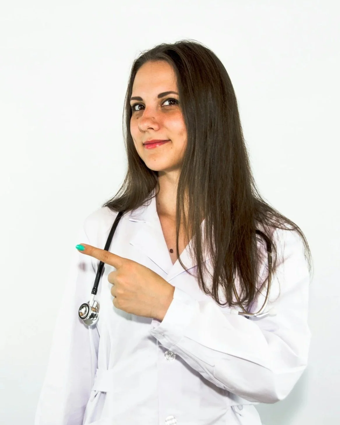 A woman in a white medical coat holding a stethoscope, pointing to the side, with a slight smile, standing against a plain white background.