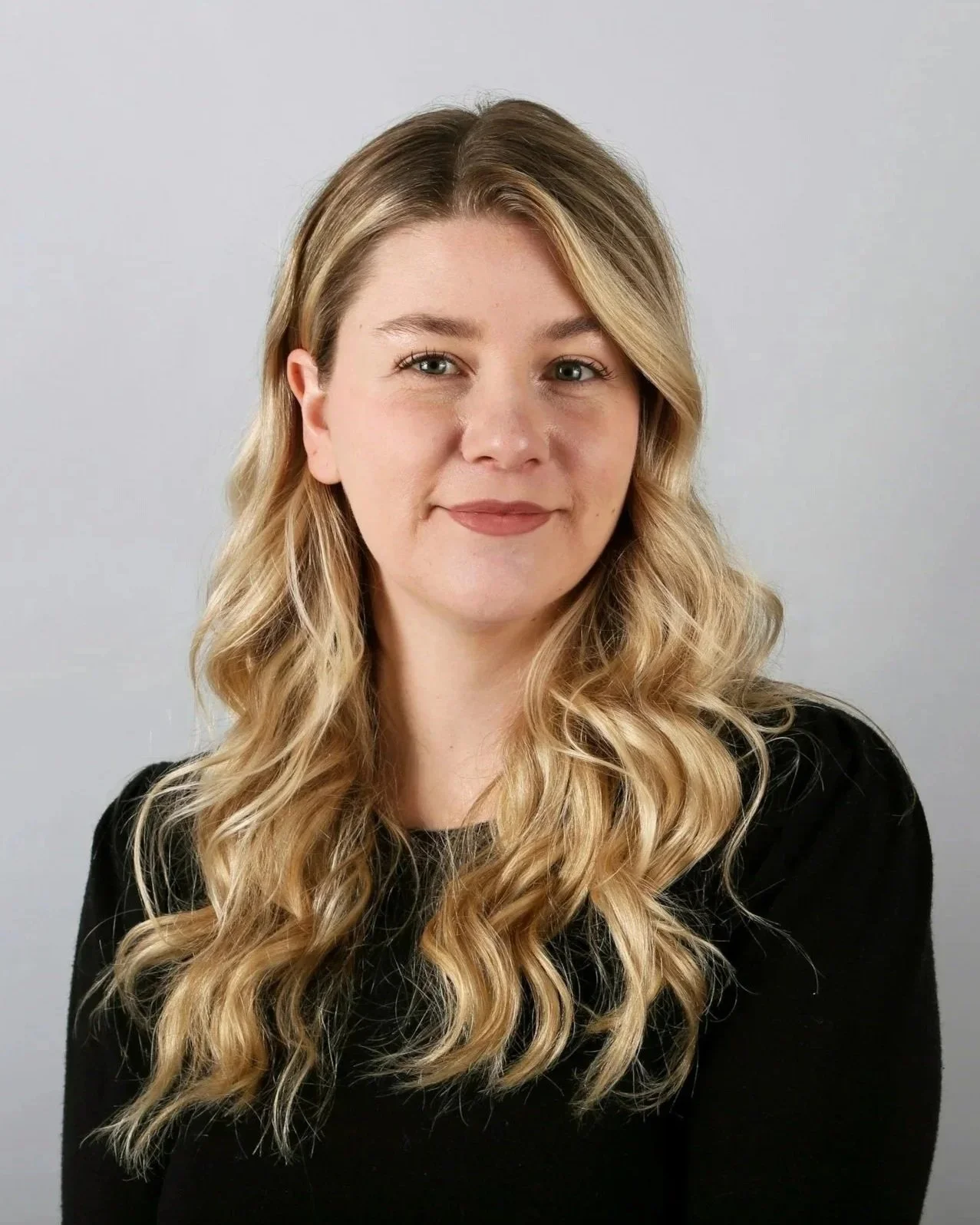 A woman with long, wavy blonde hair wearing a black top, smiling slightly, against a plain light gray background.