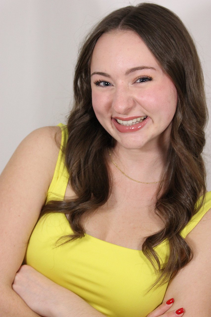 Young woman with long brown hair wearing a yellow sleeveless top, smiling with braces and red nail polish, standing against a plain wall.