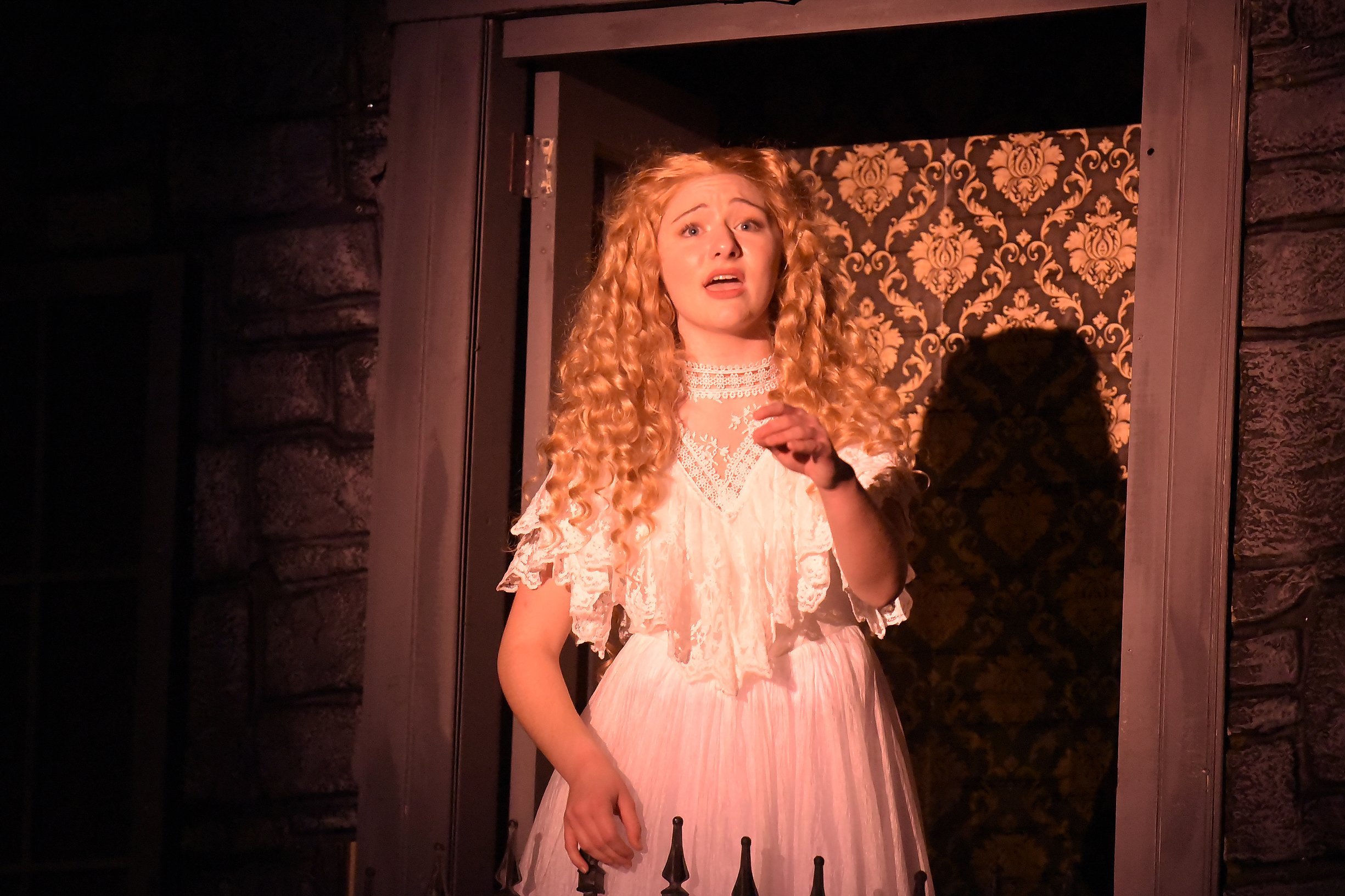 A woman in a vintage-style, white lace dress with ruffled sleeves standing behind a window or door with dark framing. She has curly red hair and a distressed or surprised facial expression, with her hand resting on her chest. The background features ornate wallpaper with a floral pattern.