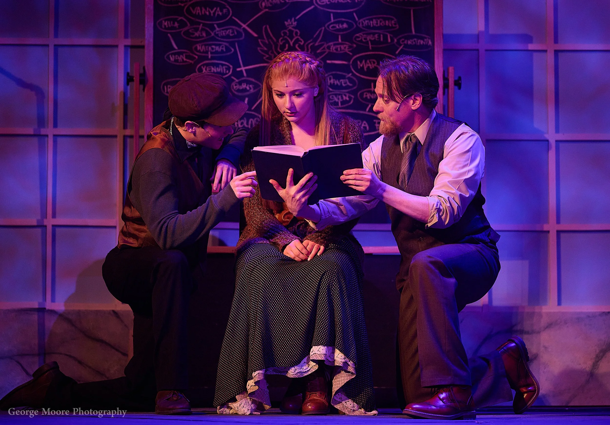 Four actors on stage, three men and one woman, engaged in a theatrical scene. The woman is seated at the center, and the men are surrounding her, looking at a book she is holding. The background features a dark chalkboard with writing and diagrams. The stage is lit with purple and pink lighting.