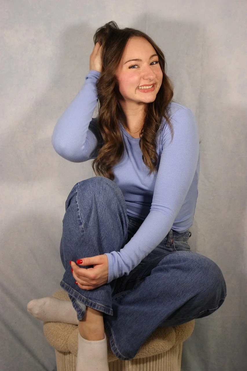 A young woman with long wavy brown hair, wearing a light blue long sleeve top and jeans, sitting cross-legged on a beige stool, smiling and playfully tugging her hair while looking at the camera, against a plain gray background.