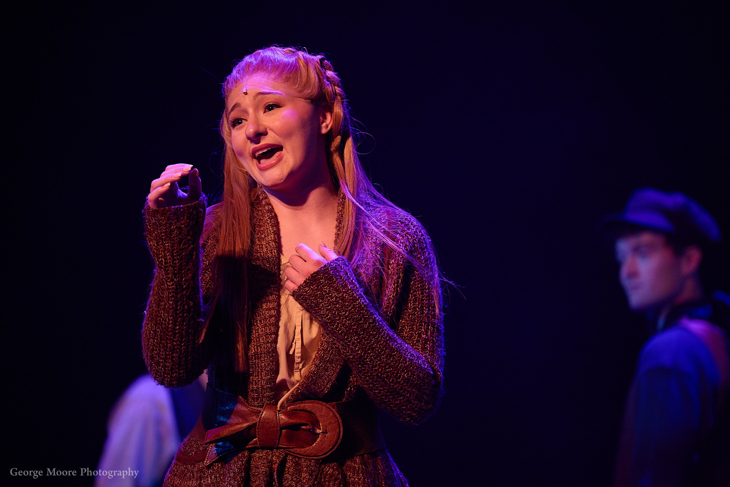 A woman with long red hair and a braid, wearing a brown textured sweater and a leather belt, appears to be singing or speaking passionately on stage, with a dark background.