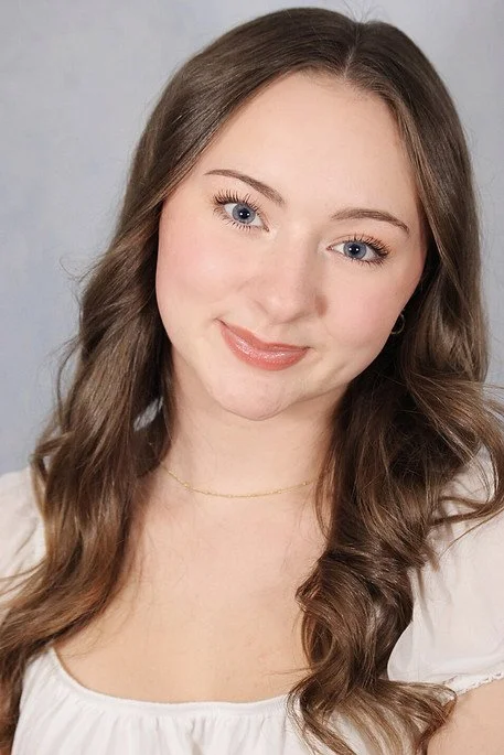 Portrait of a young woman with light skin, blue eyes, long wavy brown hair, and wearing a white top and a delicate gold necklace, smiling slightly against a gray background.