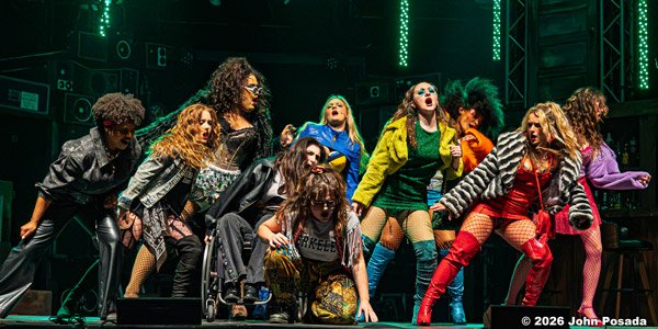 A group of women performing on stage with energetic poses, colorful outfits, and bright lighting.