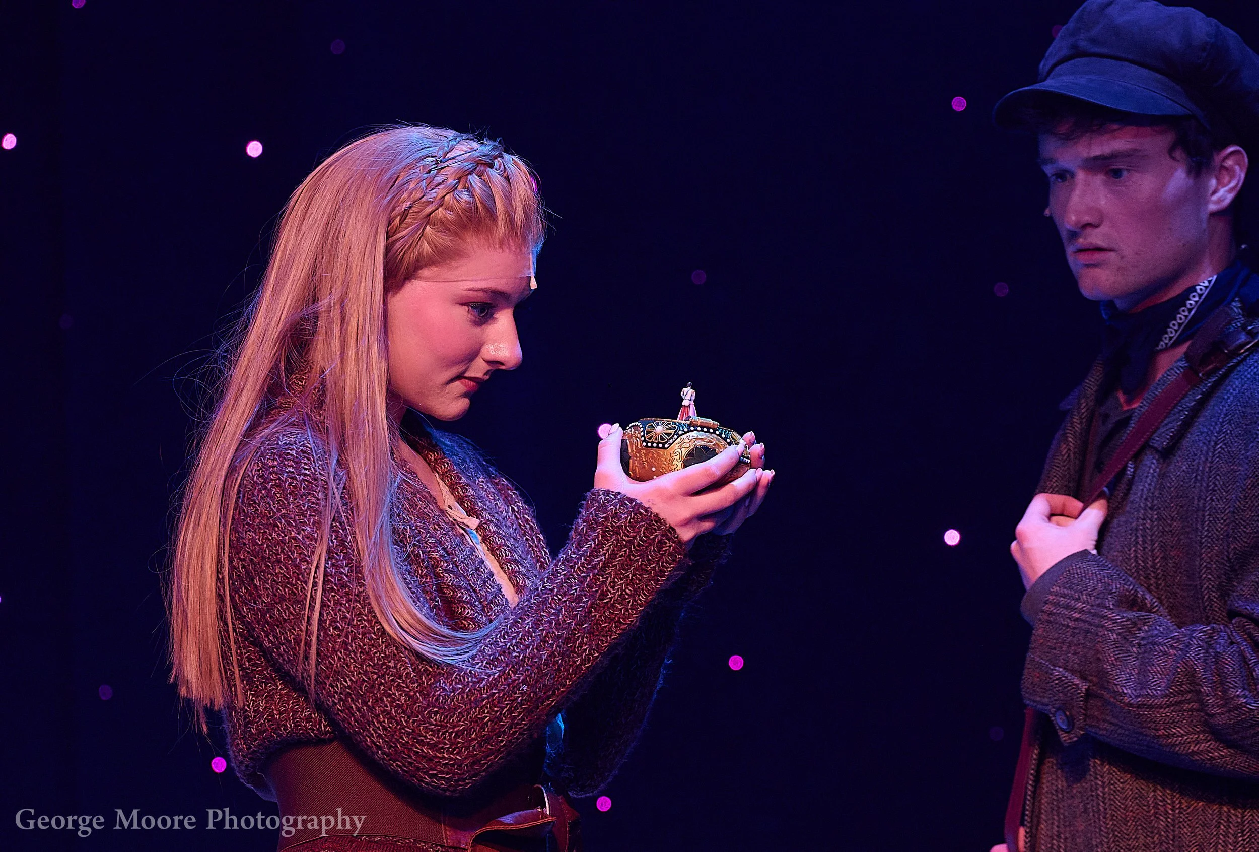A young woman with long red hair braided at the top is holding a decorative cup and looking at it. A young man with a dark cap is standing next to her looking at the cup as well, in a dark stage setting with pink and purple lighting.