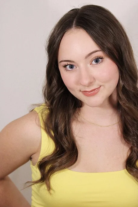 Young woman with long wavy brown hair wearing a yellow sleeveless top and a delicate gold necklace.