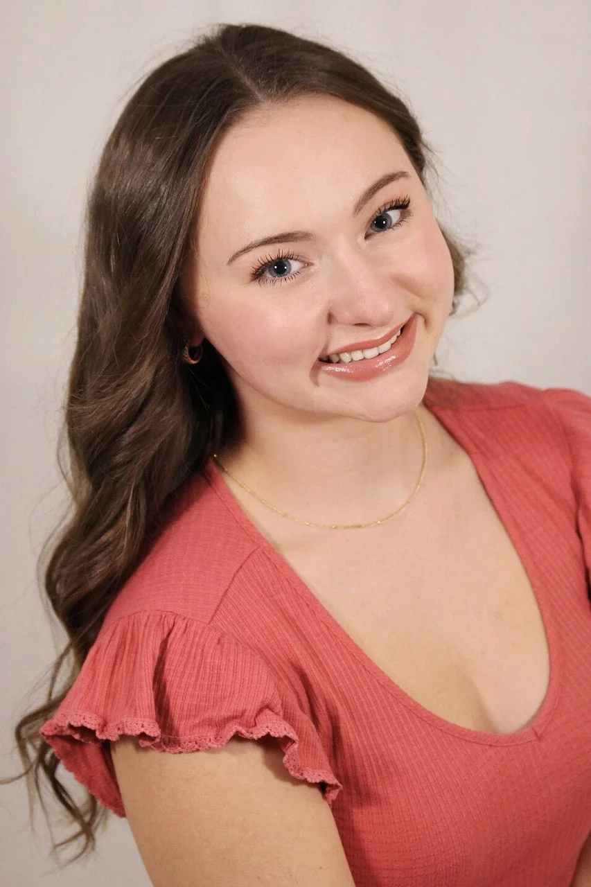 A young woman with long brown hair, blue eyes, and light skin, smiling and looking slightly to the side, wearing a pink top with ruffled sleeves and a delicate gold necklace.