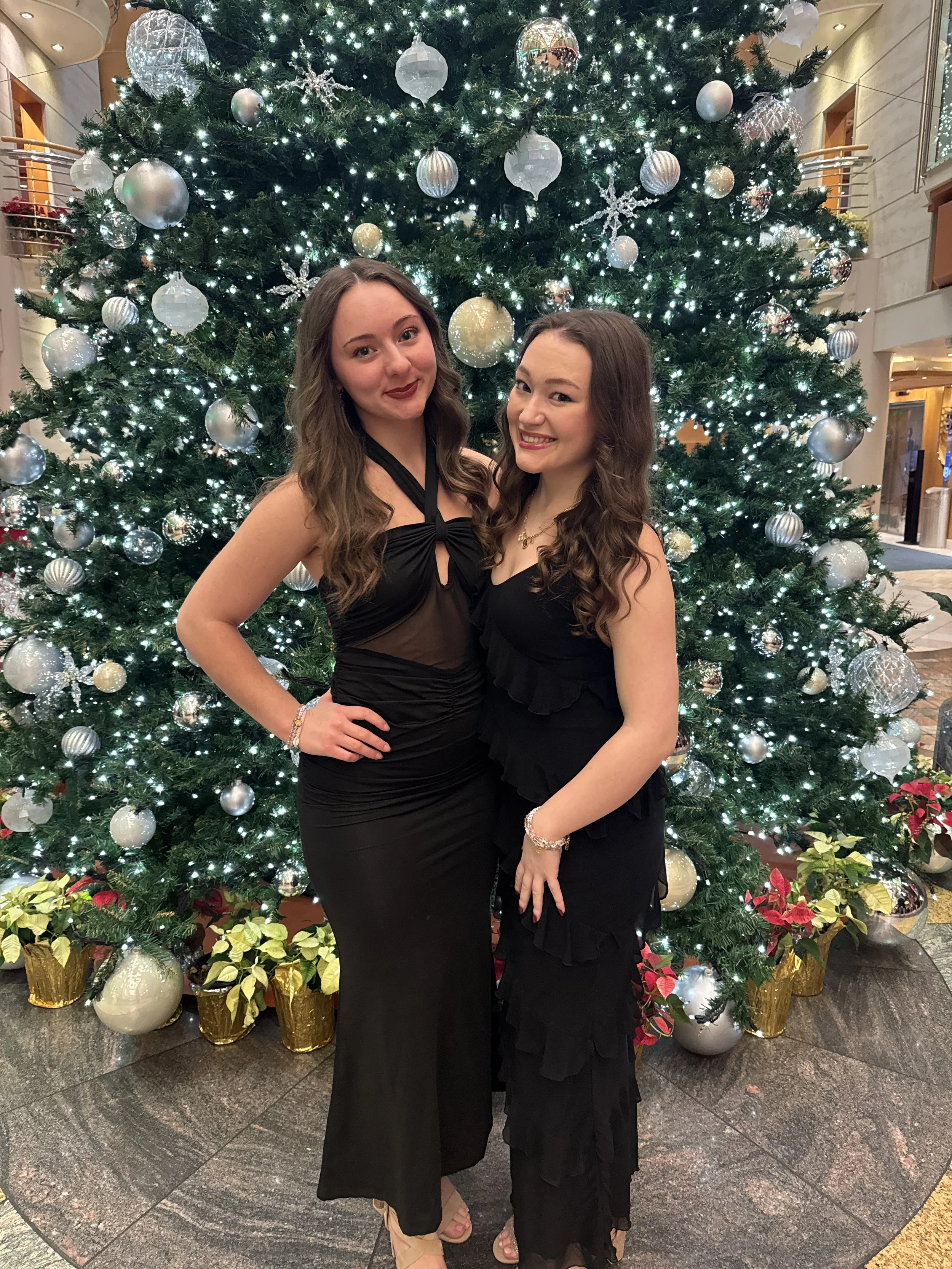 Two women in formal black dresses standing in front of a decorated Christmas tree with white and silver ornaments, surrounded by holiday plants in a hotel lobby.