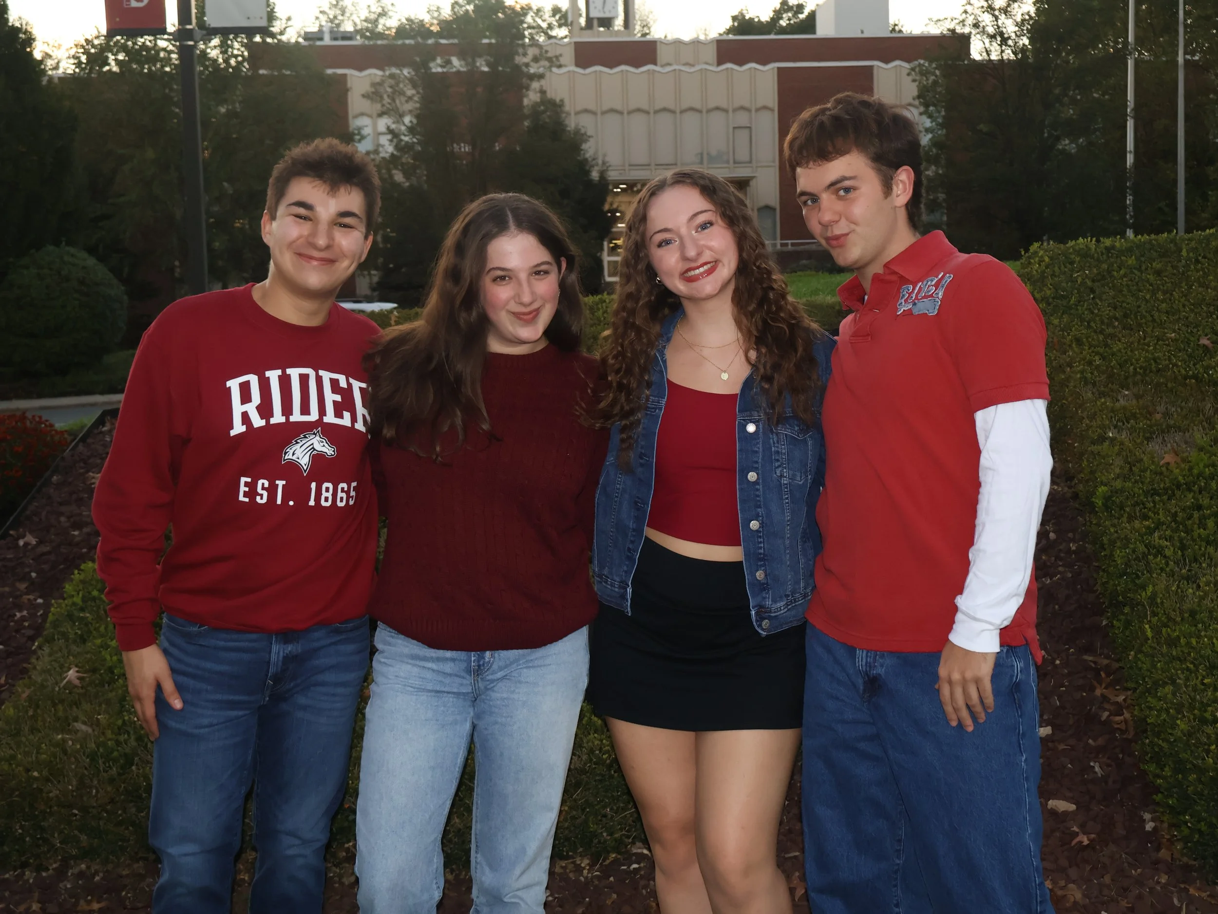 Four teenagers, two boys and two girls, standing outdoors at dusk, smiling at the camera. They are dressed casually, with the teenagers wearing red and denim clothing. The background features greenery, bushes, and a building.