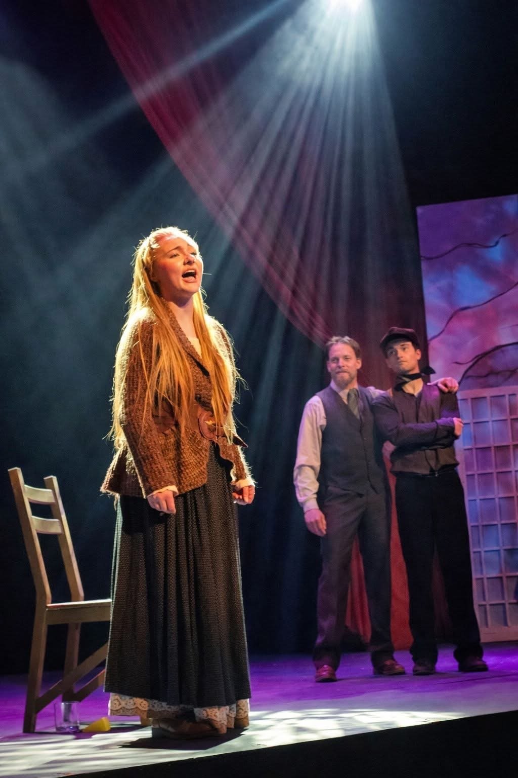 A woman with long red hair singing on stage, with two men in the background, under dramatic stage lighting, during a theatrical performance.