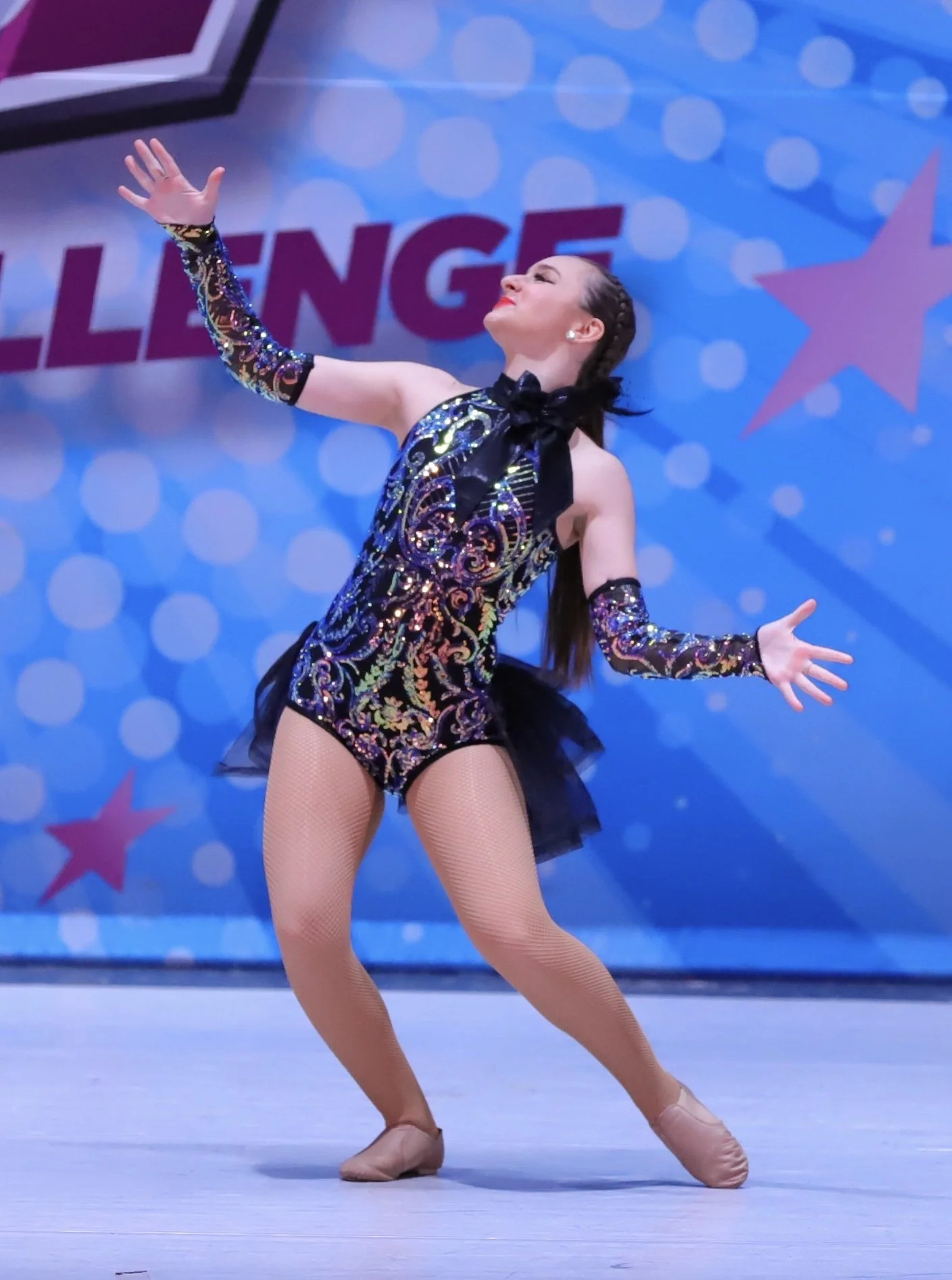 A female figure skater performing on the ice rink, wearing a sequined black dress with colorful embroidery, beige tights, and beige figure skates, with a blue background and the word 'LENGE' visible behind her.
