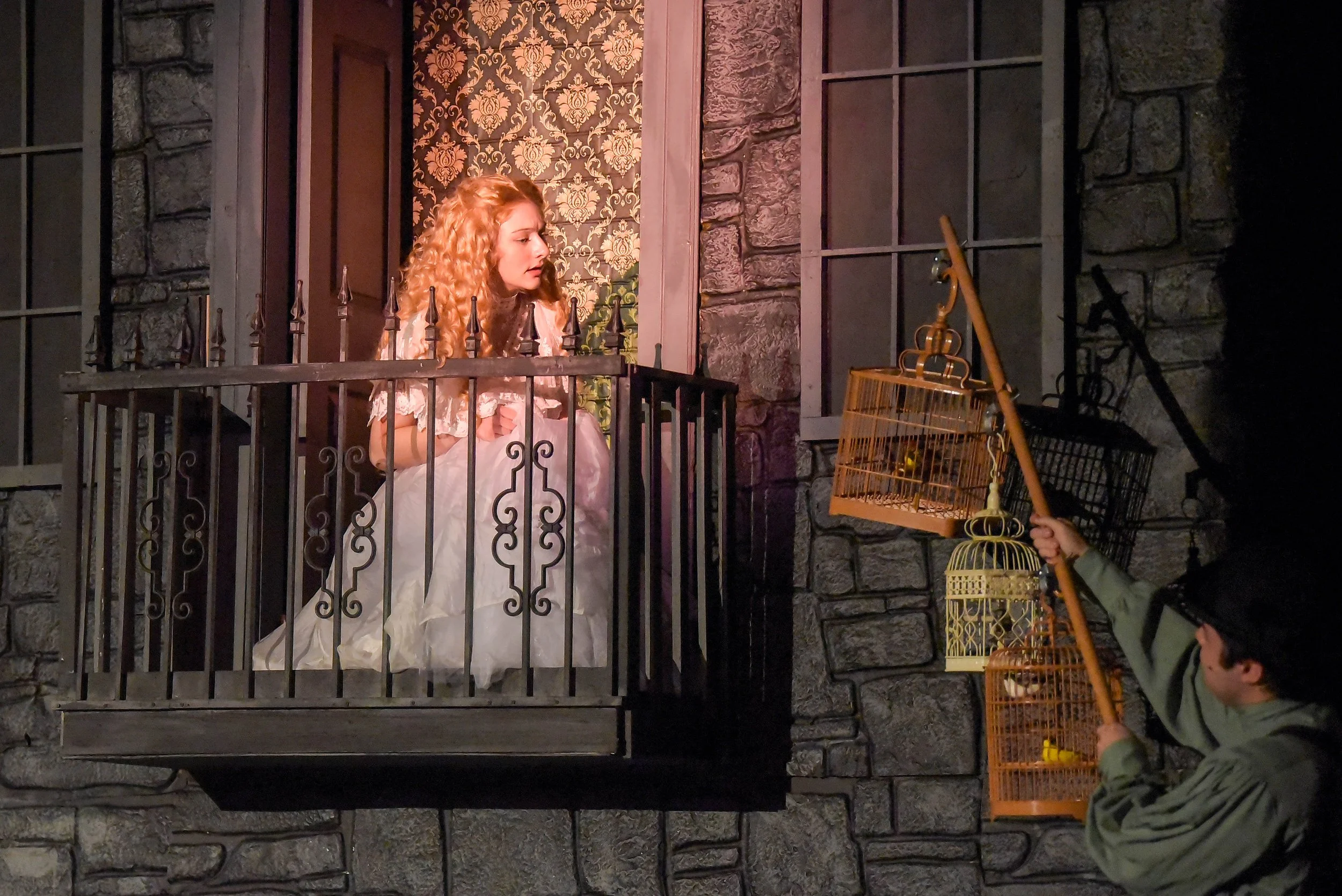 A woman dressed as a princess looking out from a small balcony with black railings, while two people hold large birdcages nearby. The scene appears theatrical or part of a performance, with textured stone wall and ornate wallpaper in the background.