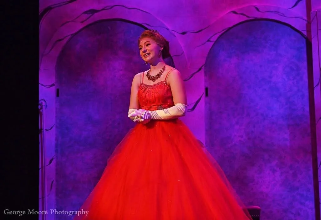 A woman dressed in a red gown with white gloves, smiling on stage with a purple background.