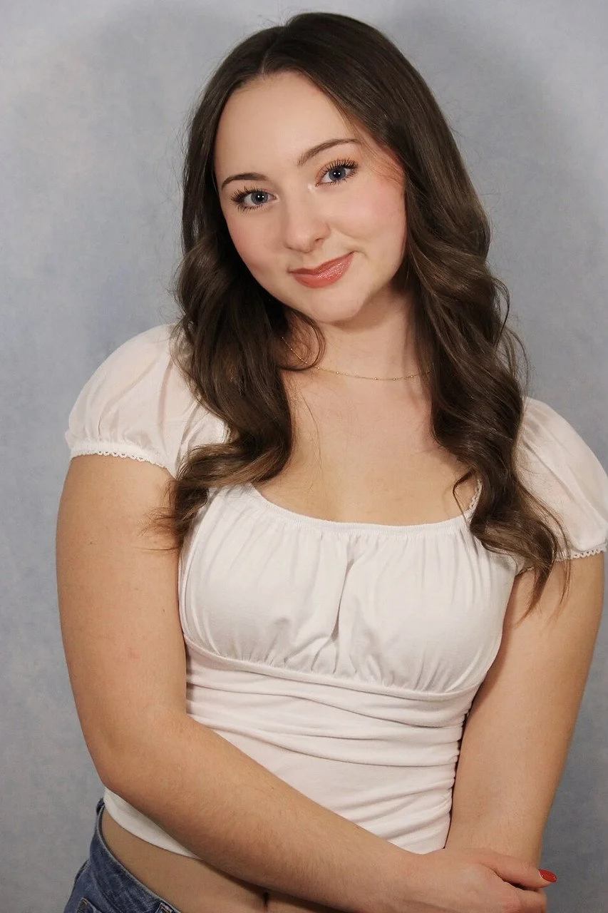 A young woman with long brown hair styled in loose waves, wearing a white puff-sleeve top and a delicate necklace, smiling softly against a plain grey background.