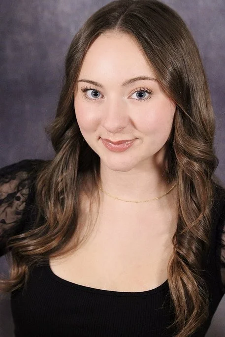 Young woman with long curly brown hair, light skin, and blue eyes, smiling softly, wearing a black top with lace sleeves and a delicate gold necklace.