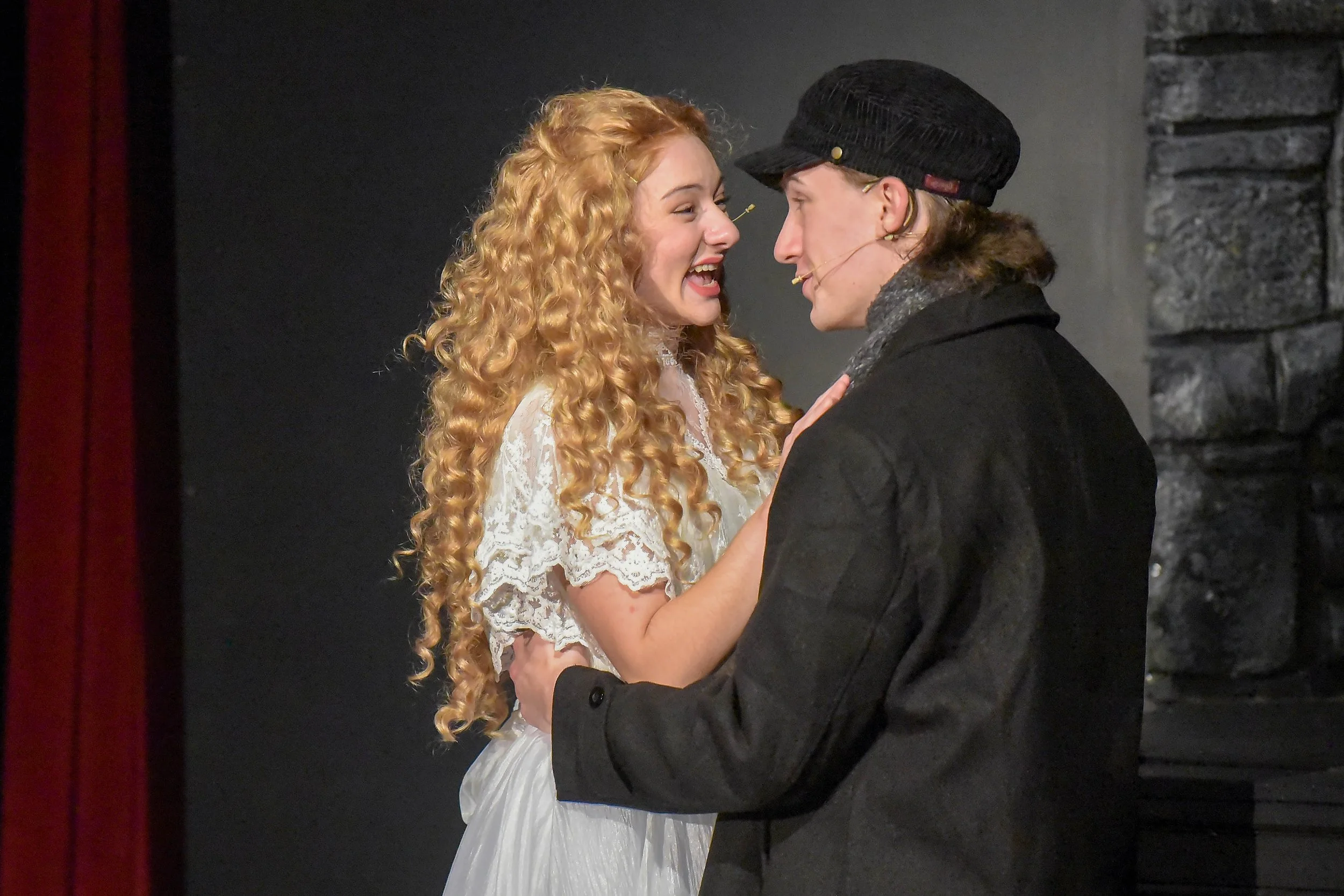 A man and woman are facing each other closely, smiling, with the woman touching the man's chest. The woman has long, curly blonde hair and is wearing a white lace dress. The man has long brown hair, a black cap, and a dark coat. They appear to be performing in a theatrical production against a dark background with stone-like wall texture.