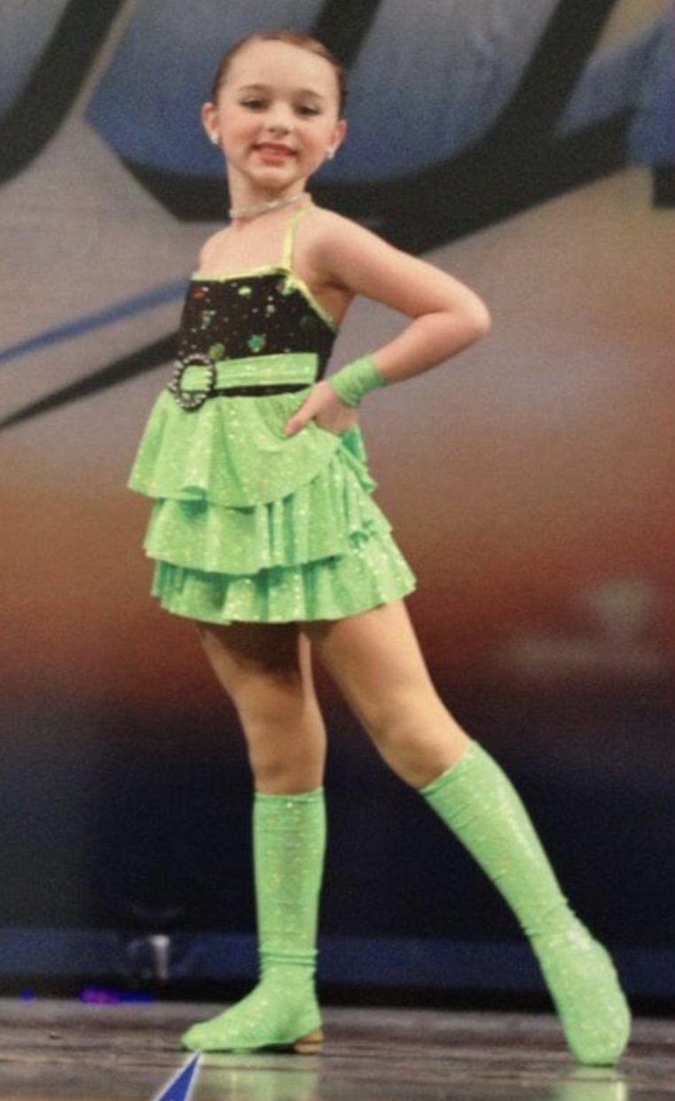 A young girl in a bright green costume with layered ruffles, matching knee-high socks, and a starry top with a belt, posing with one hand on her hip and smiling.