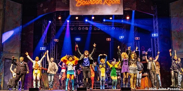 Group of performers and actors on stage at the Bourbon Room, dressed in colorful costumes, celebrating with raised hands during a theatrical production.