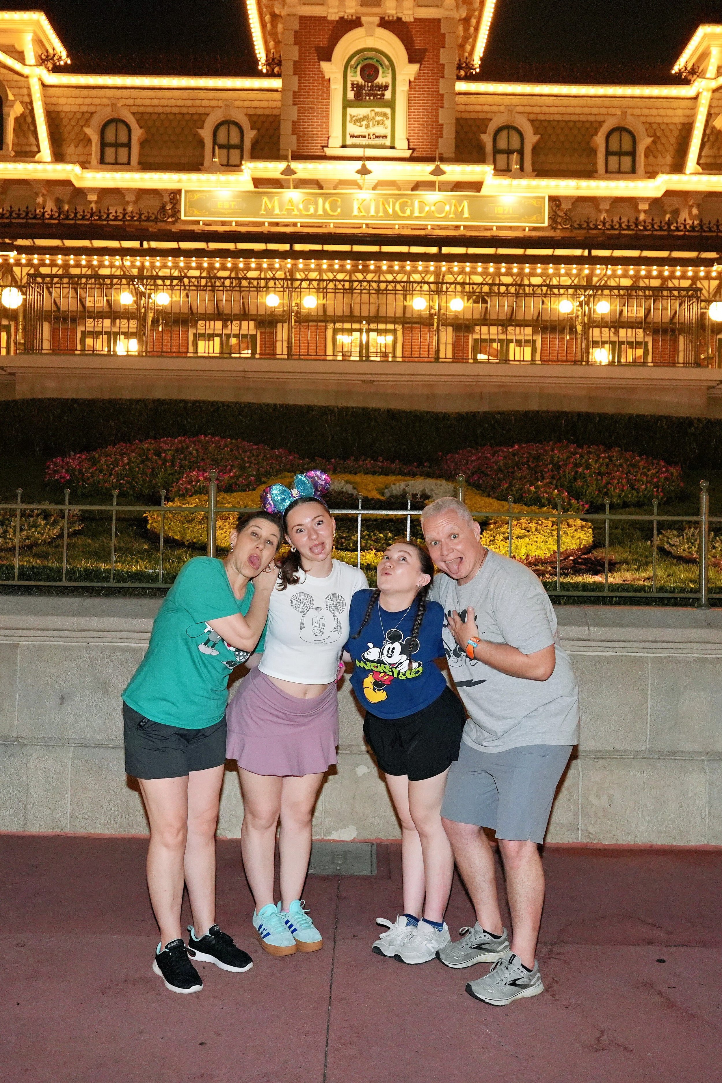 A group of four people posing in front of the Magic Kingdom entrance at night, with a well-lit, decorated building and flowers in the background.