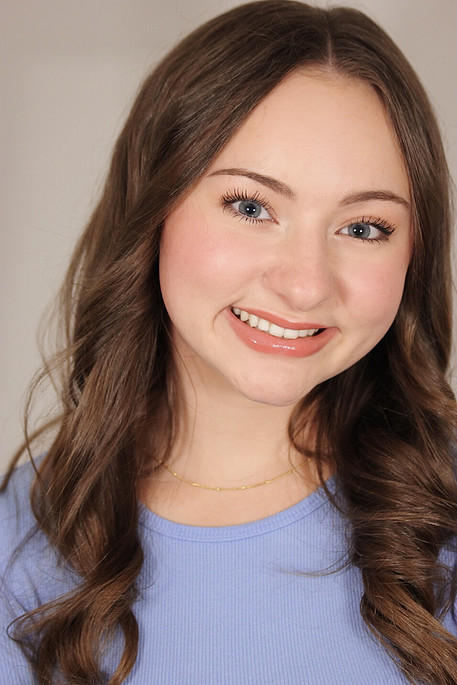 A young woman with long brown hair, blue eyes, and a smile wearing a light blue shirt and a gold necklace.