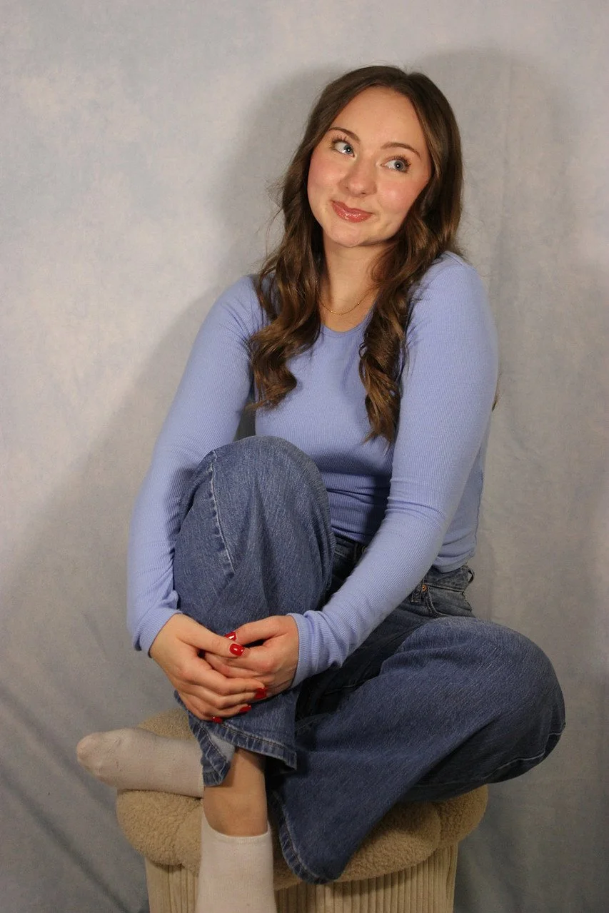 A young woman with long brown hair sitting on a small stool, wearing a light purple long sleeve shirt and blue jeans, with her legs crossed and hands clasped around one knee, smiling slightly with her head tilted to the side.