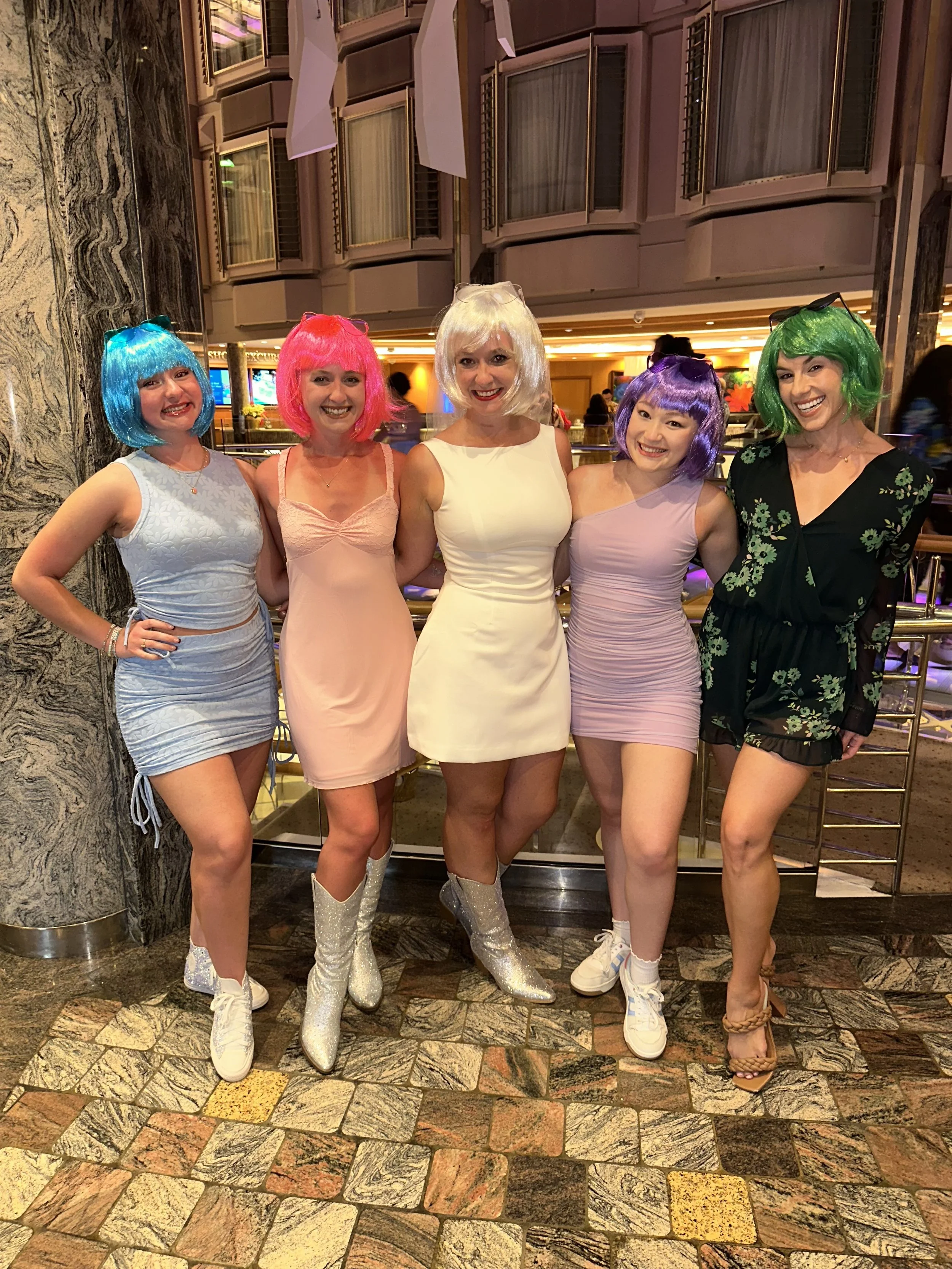 Five women dressed in colorful wigs and playful outfits standing together in a hotel lobby or casino area, smiling for the camera.