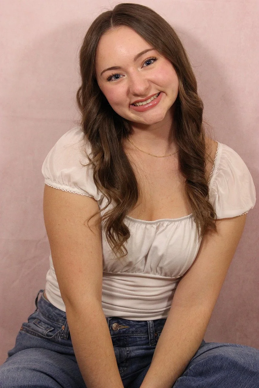 A young woman with long brown hair, smiling, wearing a white blouse and blue jeans, sitting against a pink background.