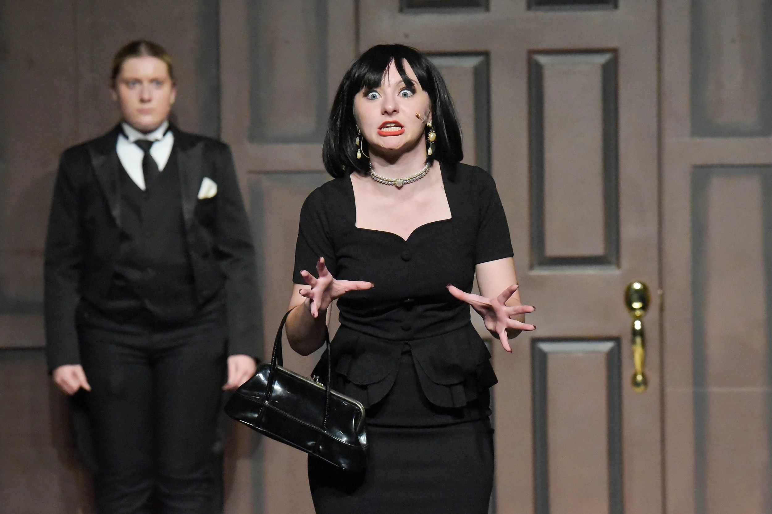 A woman with black hair and bright blue eyes, wearing a black dress and jewelry, is speaking with animated hand gestures. She has a black handbag hanging from her left arm. Behind her, a person in a black suit with a white shirt and black tie stands quietly. The scene appears to be indoors with a brown door in the background.