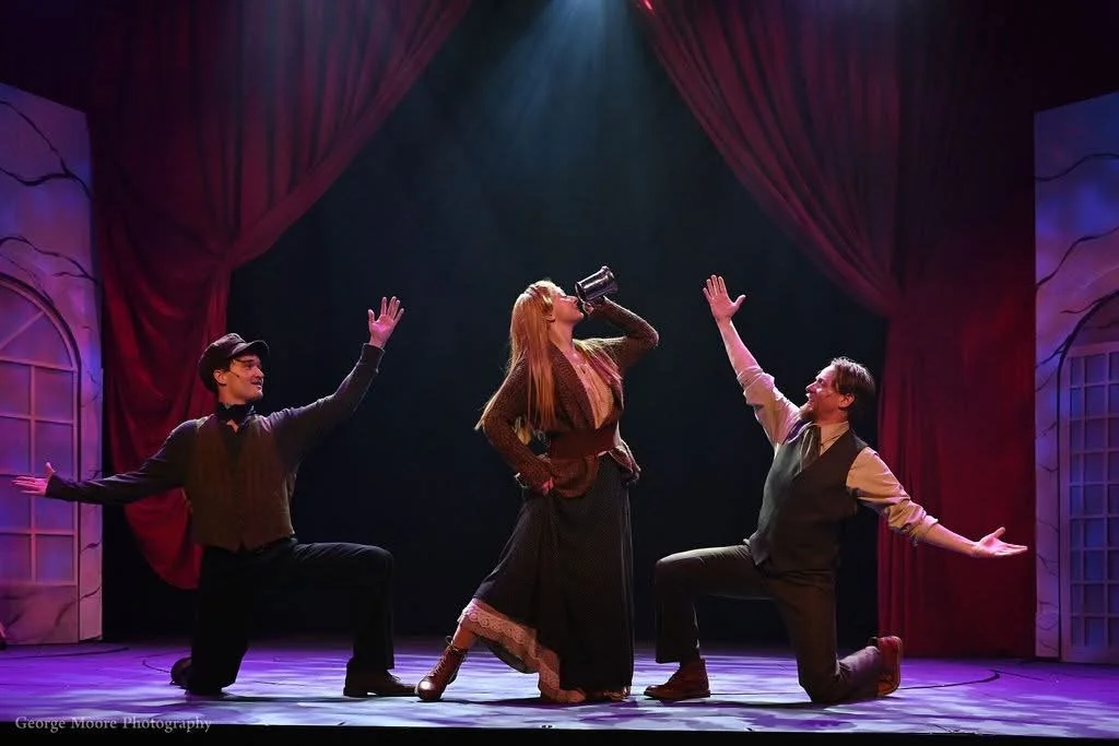 Three actors performing on stage with red curtains, two men kneeling and woman standing, one man raising hand, woman drinking from a mug.