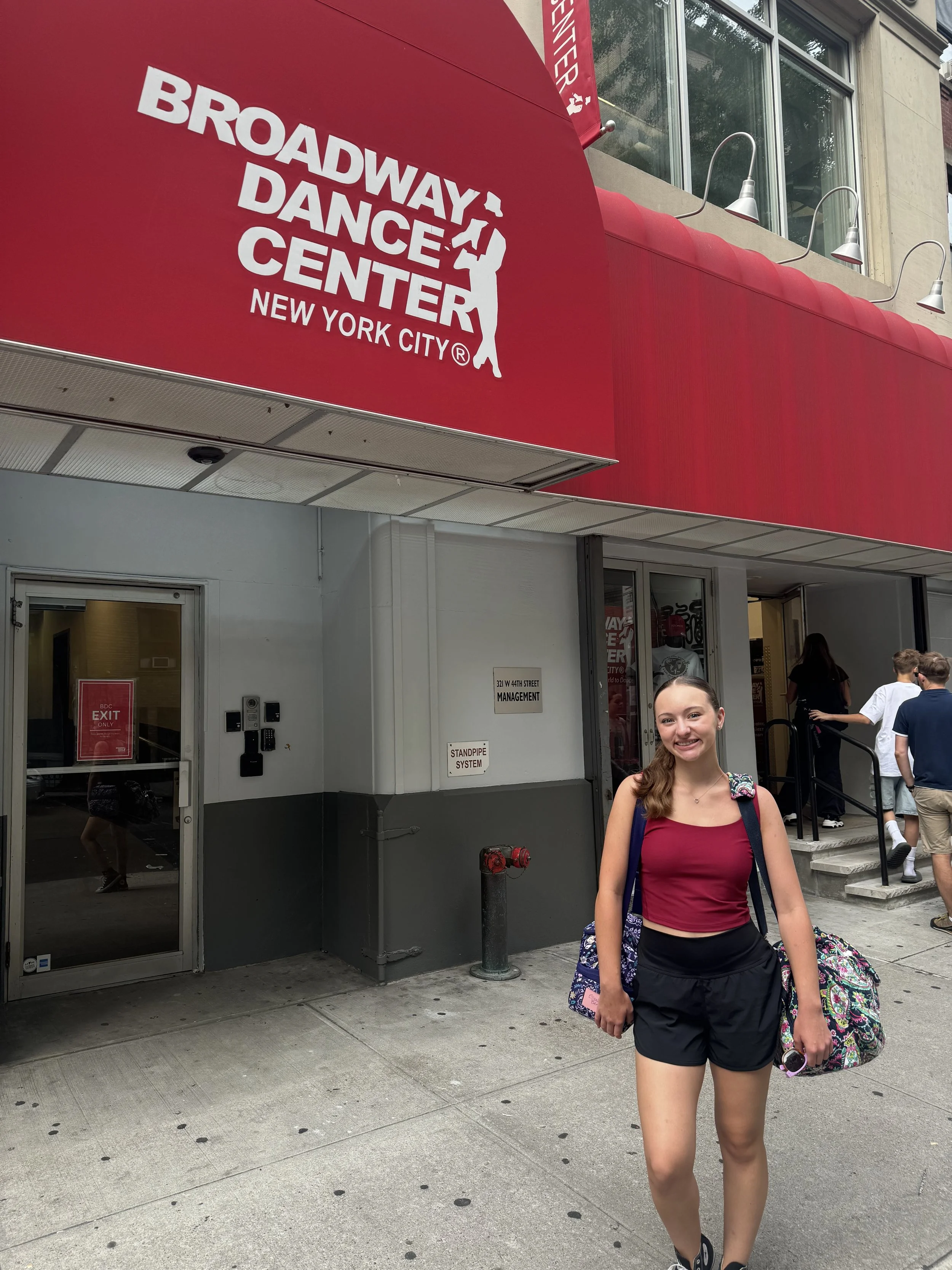 A smiling young woman with long hair, wearing a sleeveless burgundy top and black shorts, stands outside the Broadway Dance Center in New York City. She carries a colorful bag over her shoulder and holds a pink wallet or phone in her hand. Other people are visible entering or exiting the building behind her.