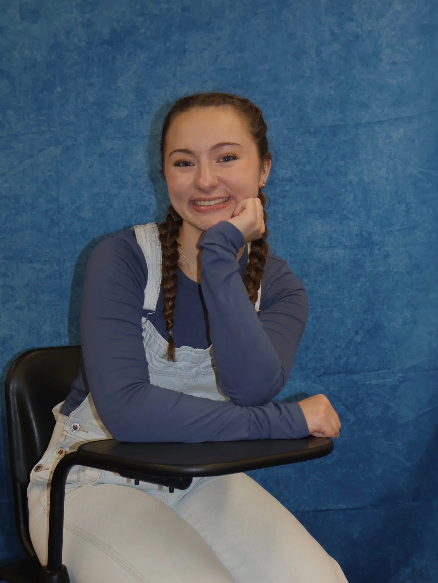 A young woman with braided hair sitting on a black chair, smiling, with her chin resting on her hand, against a blue wall background.