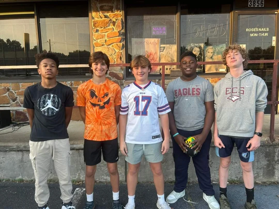 Five boys standing in front of a storefront with large windows and stone accents, posing for a group photo outside during daytime.