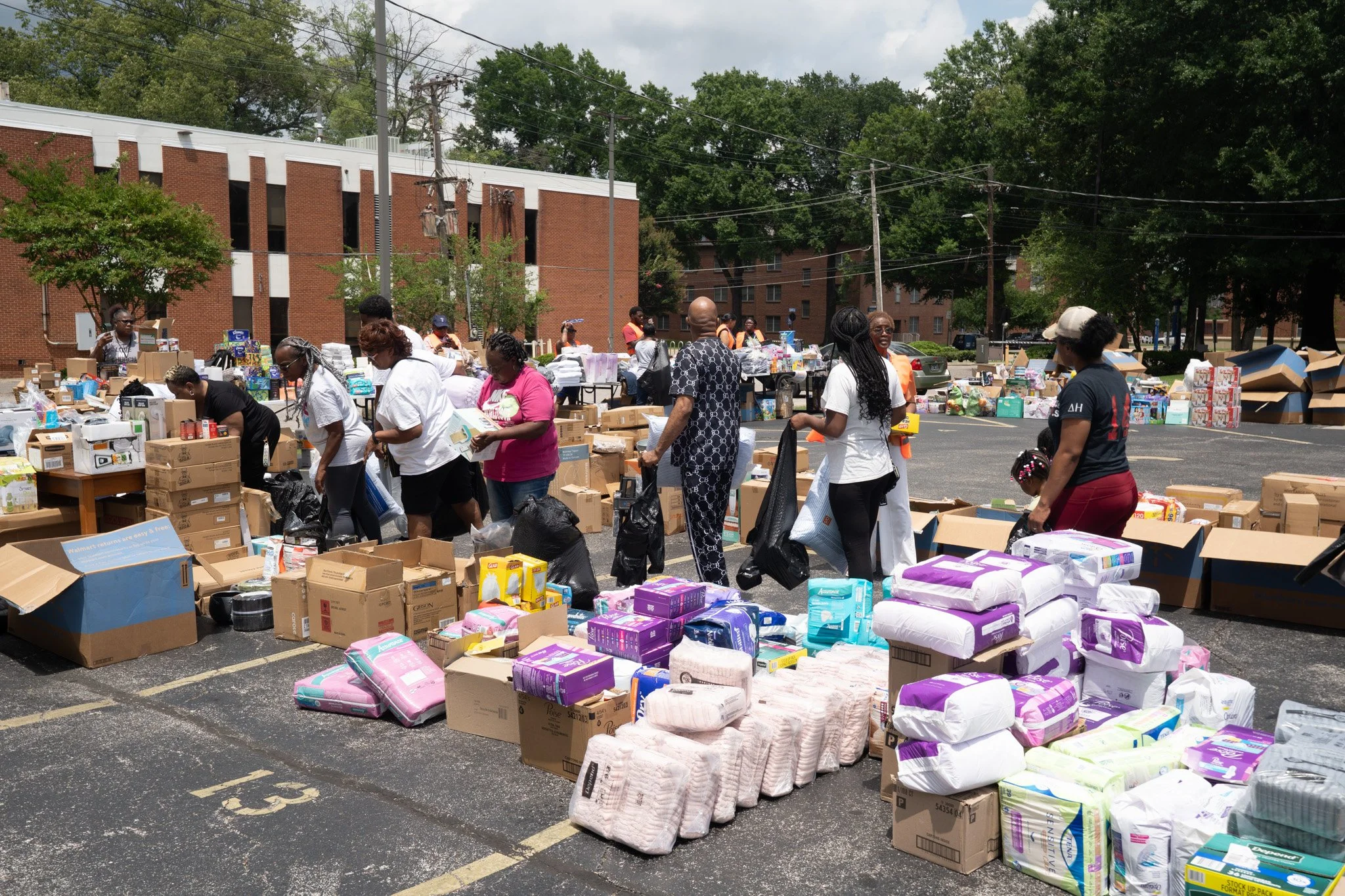 People at a carport sale with tables and piles of diapers, baby supplies, and boxed items in a parking lot with trees and a brick building in the background.