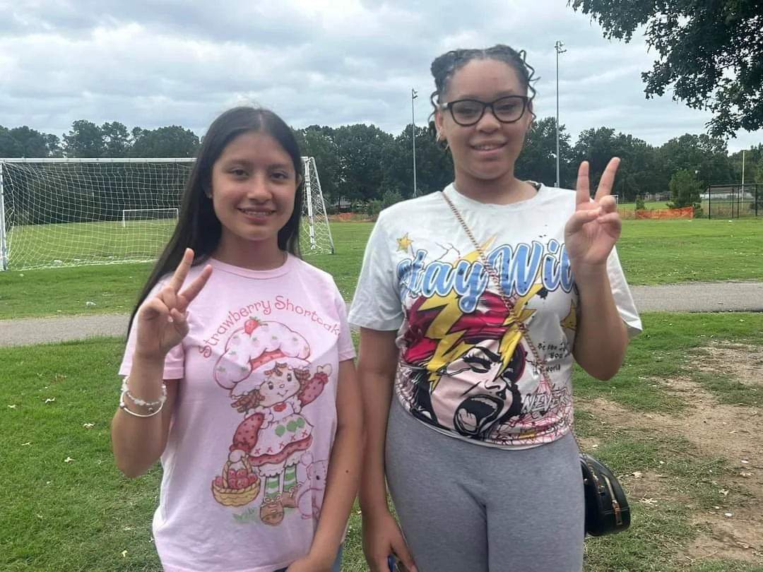 Two young girls standing outdoors on a grassy field with soccer goals in the background, both making peace signs with their hands and smiling at the camera.