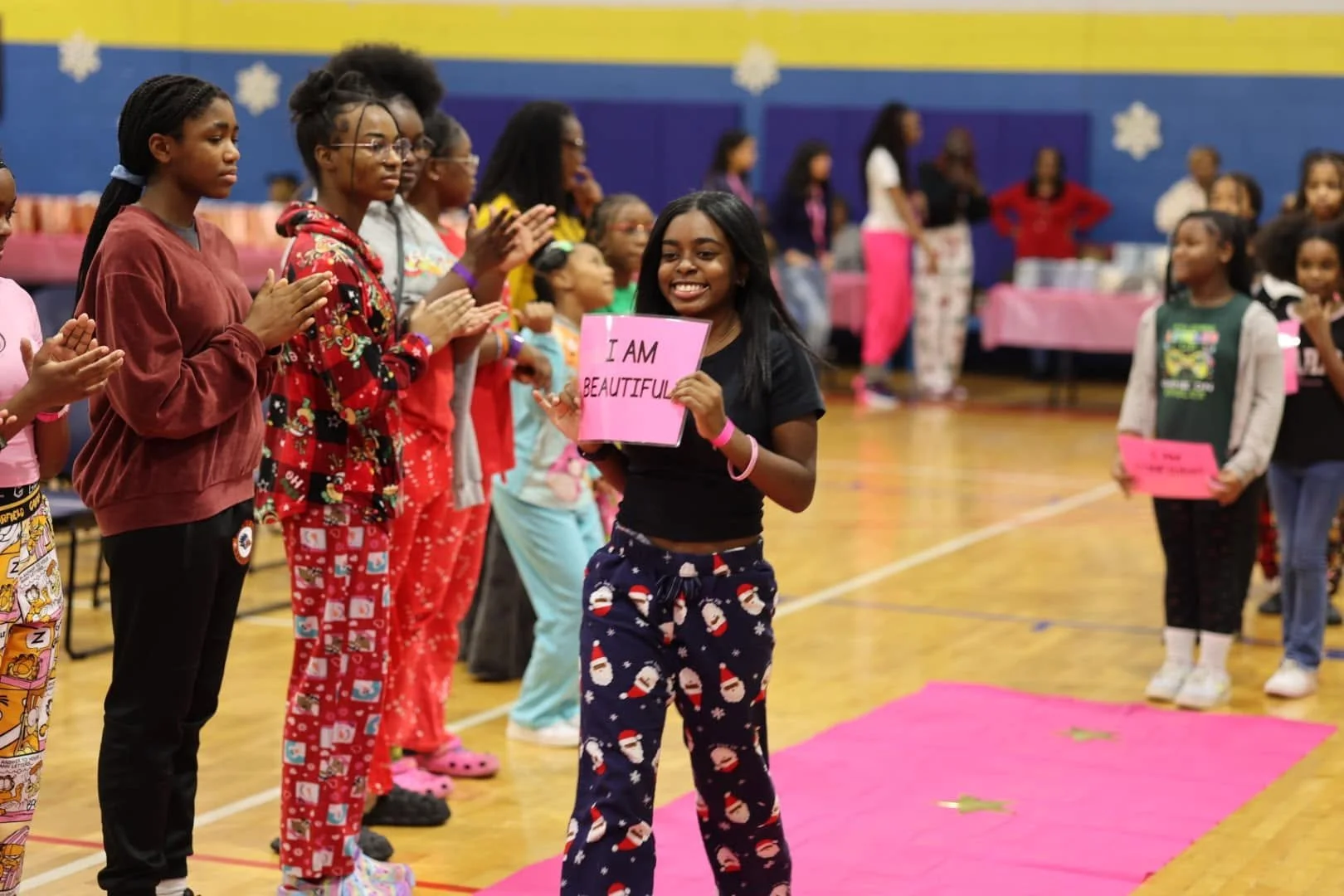 A group of children in pajamas standing in a gymnasium, some clapping and some holding signs, participating in a celebration or event. A girl in the center is holding a pink sign that reads 'I AM BEAUTIFUL' and is smiling. The decorations and pink ribbon suggest a festive or awareness event, possibly related to self-esteem or body positivity.