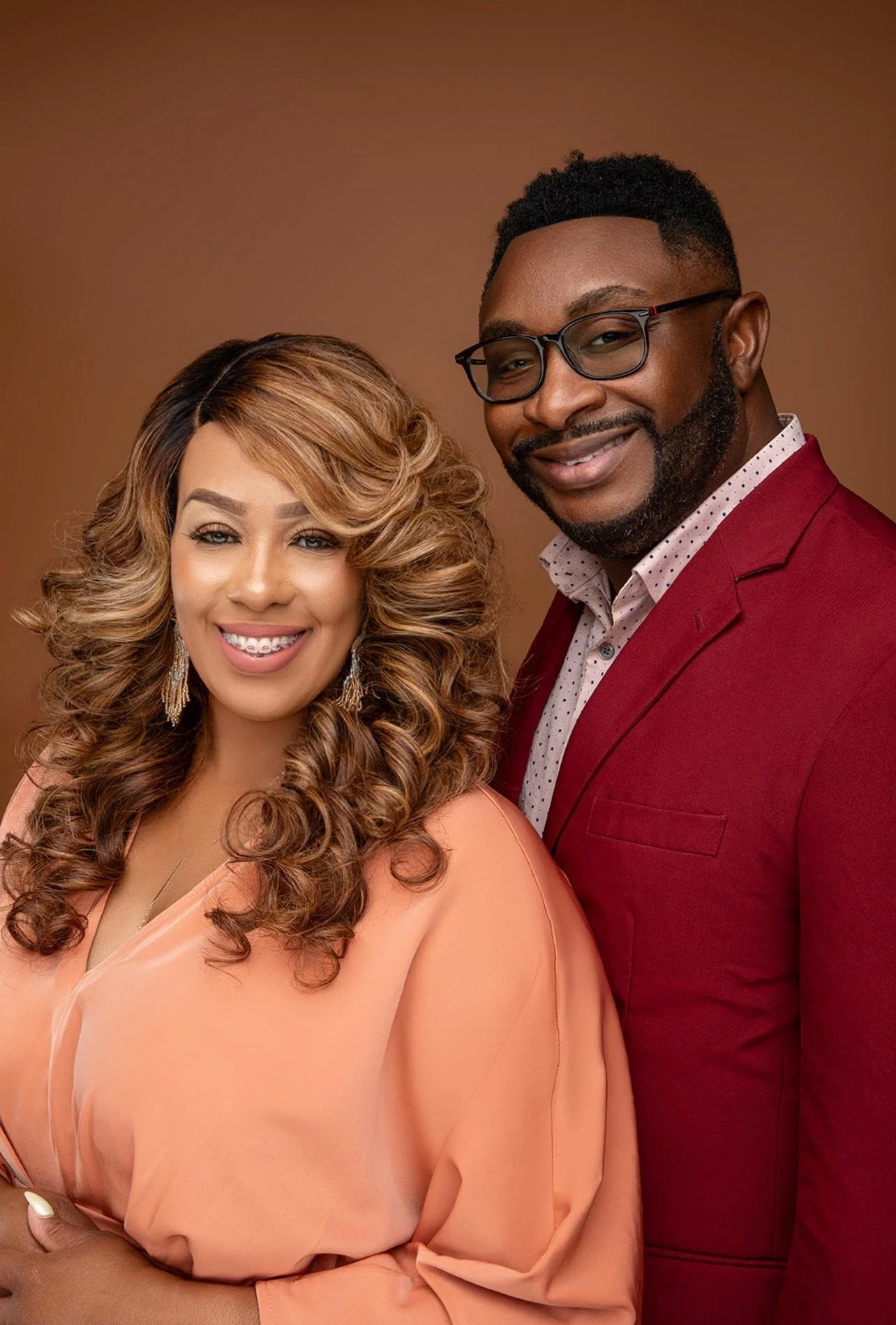 A smiling woman with long curly hair wearing a peach-colored dress and gold earrings standing next to a smiling man with glasses, a beard, wearing a red blazer and a white dotted shirt, against a brown background.