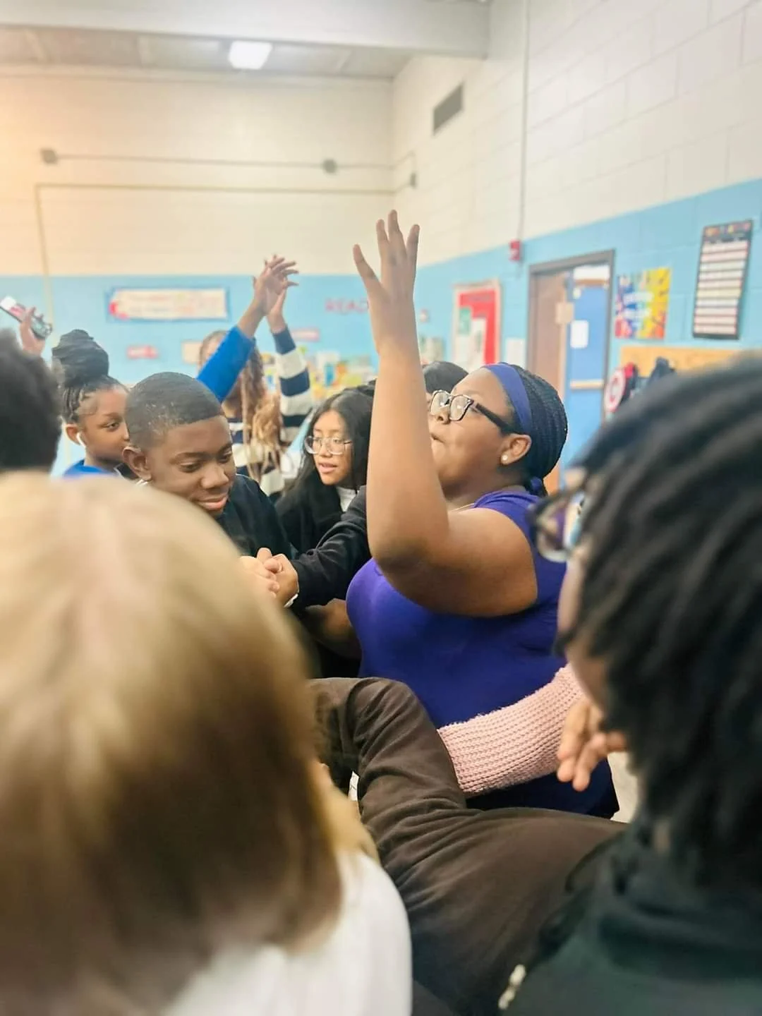 A group of students and a woman, possibly a teacher, are gathered in a school hallway. The woman, wearing glasses and a blue shirt, has her hand raised and appears to be participating in an activity with the students.