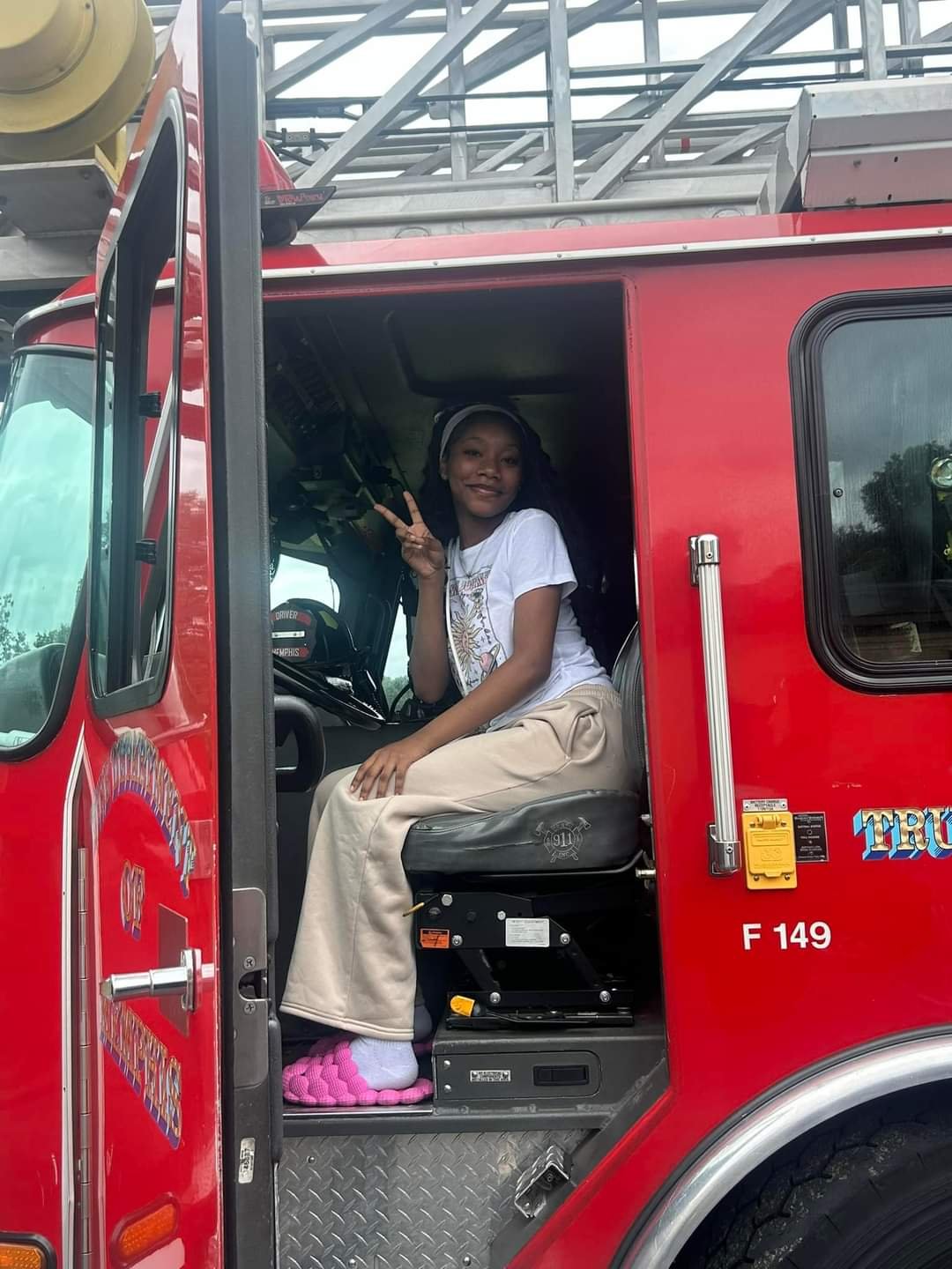 A girl smiling and making a peace sign while sitting in the driver's seat of a red fire truck.