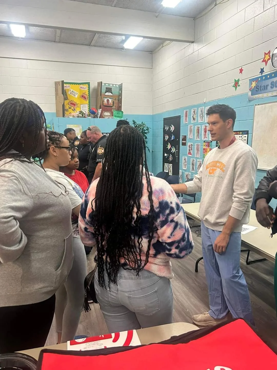 A man in a gray sweatshirt and light blue pants talking to a group of young women in a classroom with colorful wall decorations.