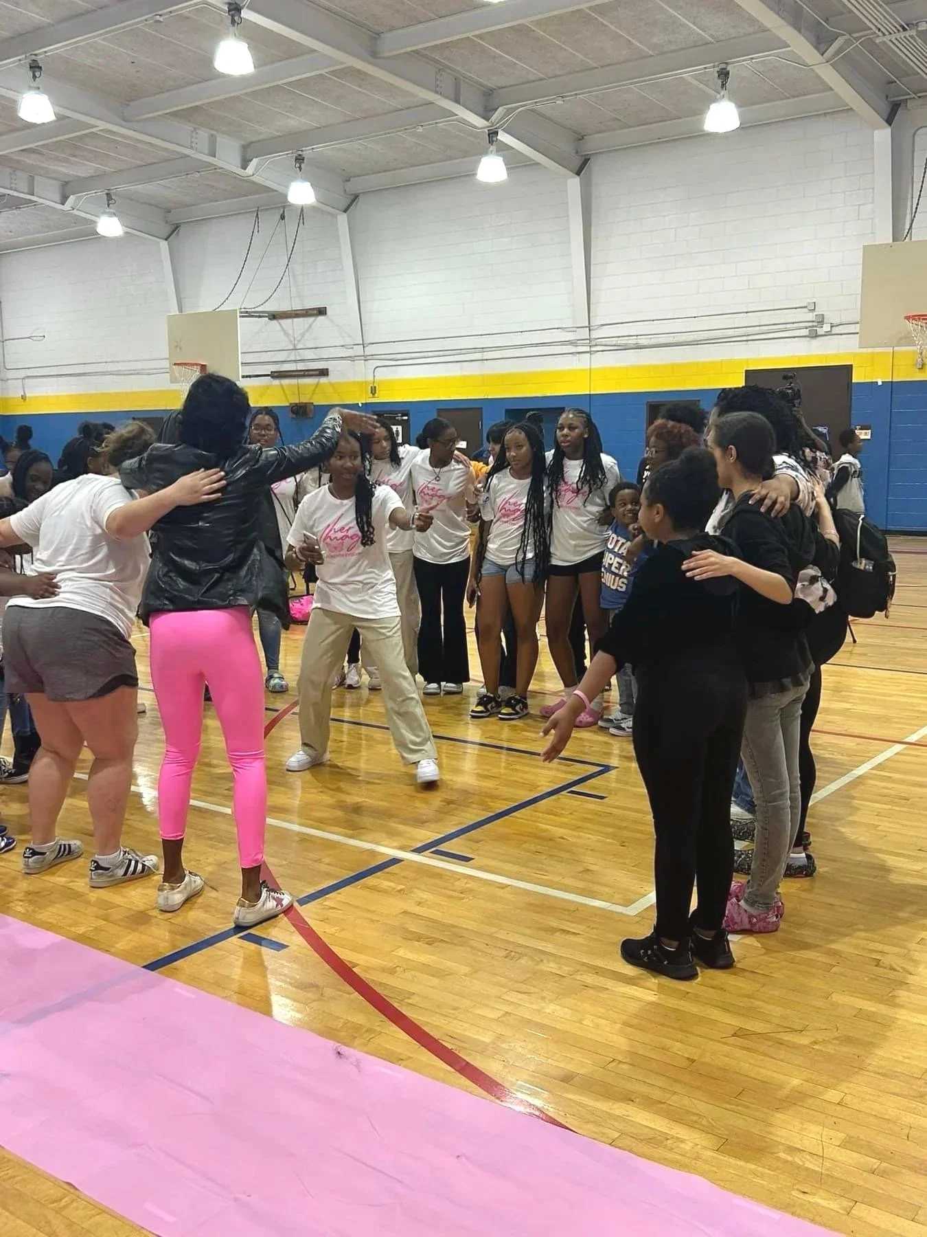 A group of diverse women and girls standing in a gymnasium, some hugging and others talking, during a community or fitness event.