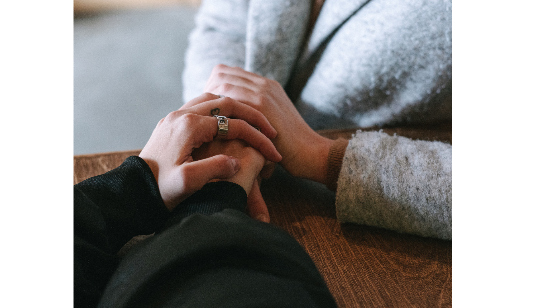 Two people holding hands on a wooden table, one person wearing a black long sleeve shirt with a ring on their finger, and the other wearing a gray sweater.